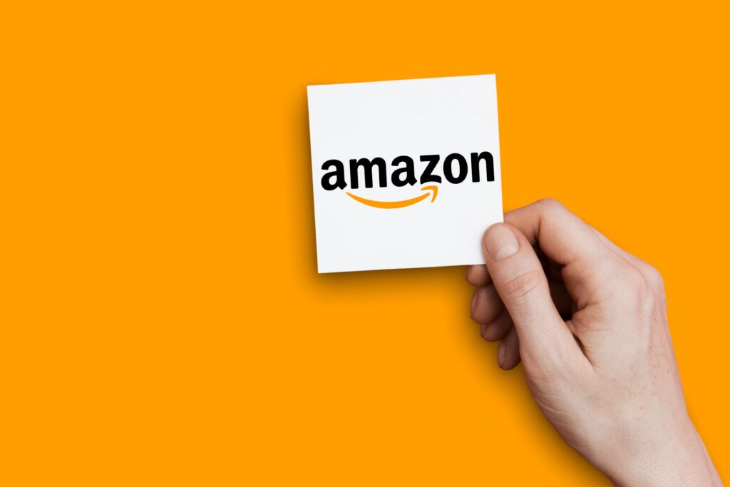 Hand hält einen weißen Zettel mit dem Amazon-Logo vor orangefarbenem Hintergrund – E-Commerce-Marktplatz und Branding-Strategie